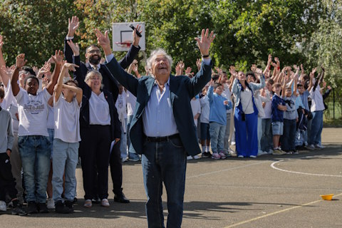 Pierre Perret levant les bras debout avec des enfants dans la cours du collège qui porte son nom.